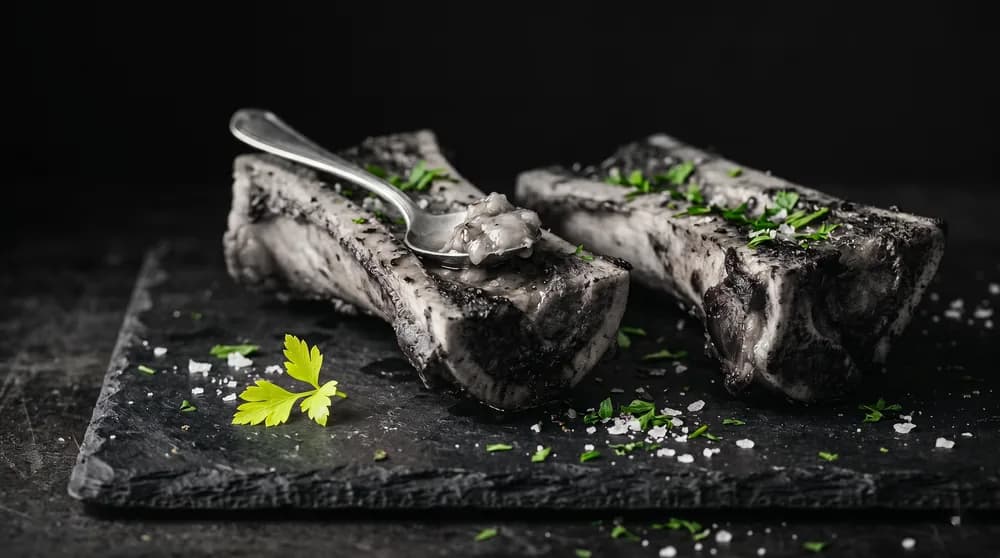 Halved marrow bones with a spoon, coarse sea salt and parsley on a dark slate board
